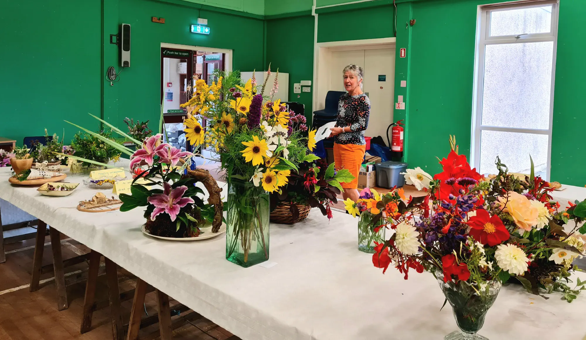 The table in the floral section, filled with flower arrangements, and Caroline in the background looking at them